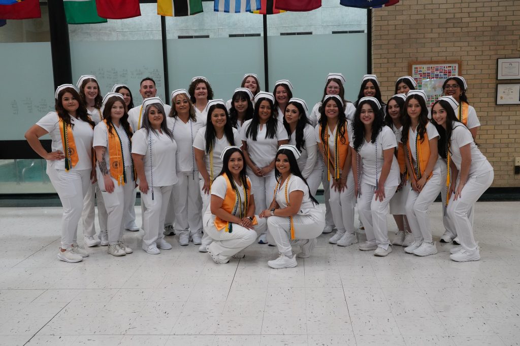 A group of students dressed in white medical uniforms and caps, posing together for a photo indoors, with international flags hanging in the background.