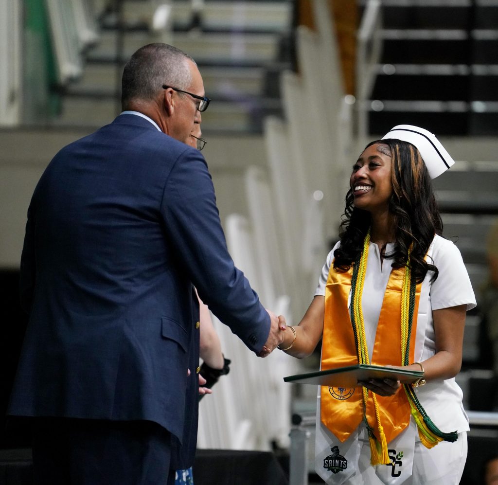 A smiling graduate in a white outfit and a graduation cap shakes hands with a man in a suit, holding a diploma, during a graduation ceremony.